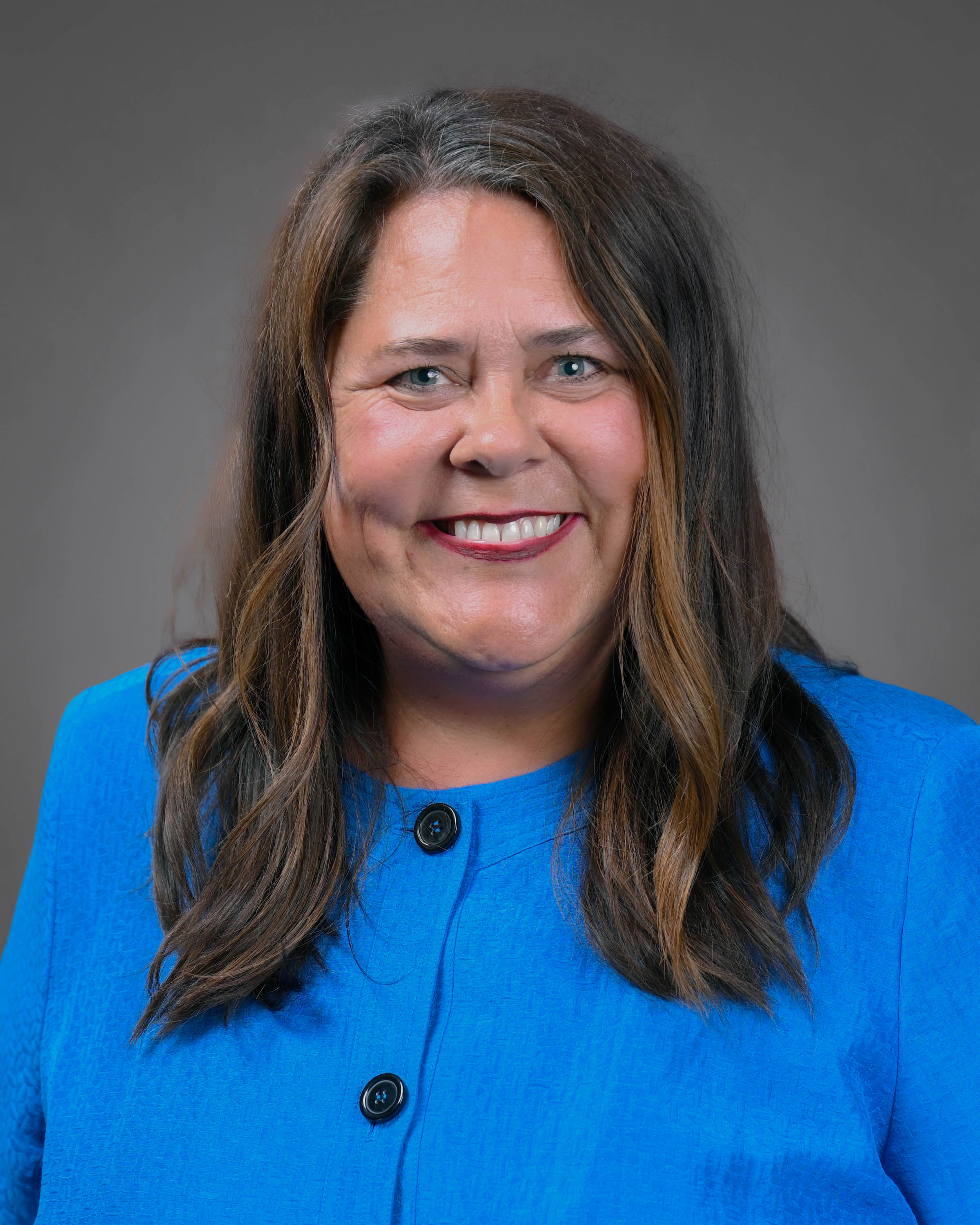 Professional headshot of a smiling woman with long dark hair, wearing a bright blue jacket against a gray studio background.